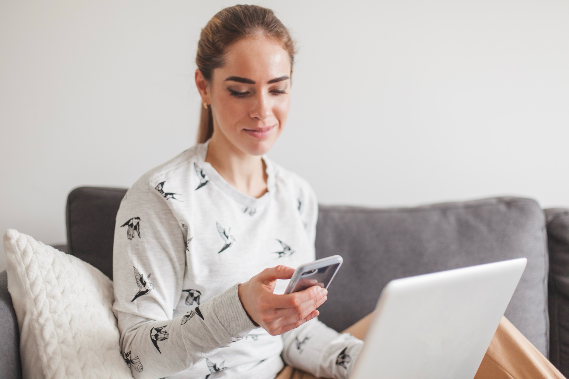 Kvinna i soffan tittar i sin mobiltelefon med en bärbar dator i knät